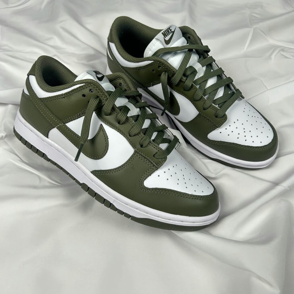 Stylish Nike Dunk Low sneakers in olive and white placed on a soft white fabric background.